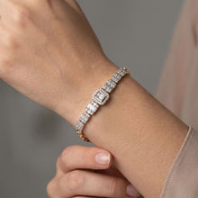 Load image into Gallery viewer, A close-up of a delicate bracelet worn on a wrist, featuring a row of sparkling clear rectangular gemstones in a yellow gold setting. The background is a soft, blurred gray with a hint of a light pink garment.
